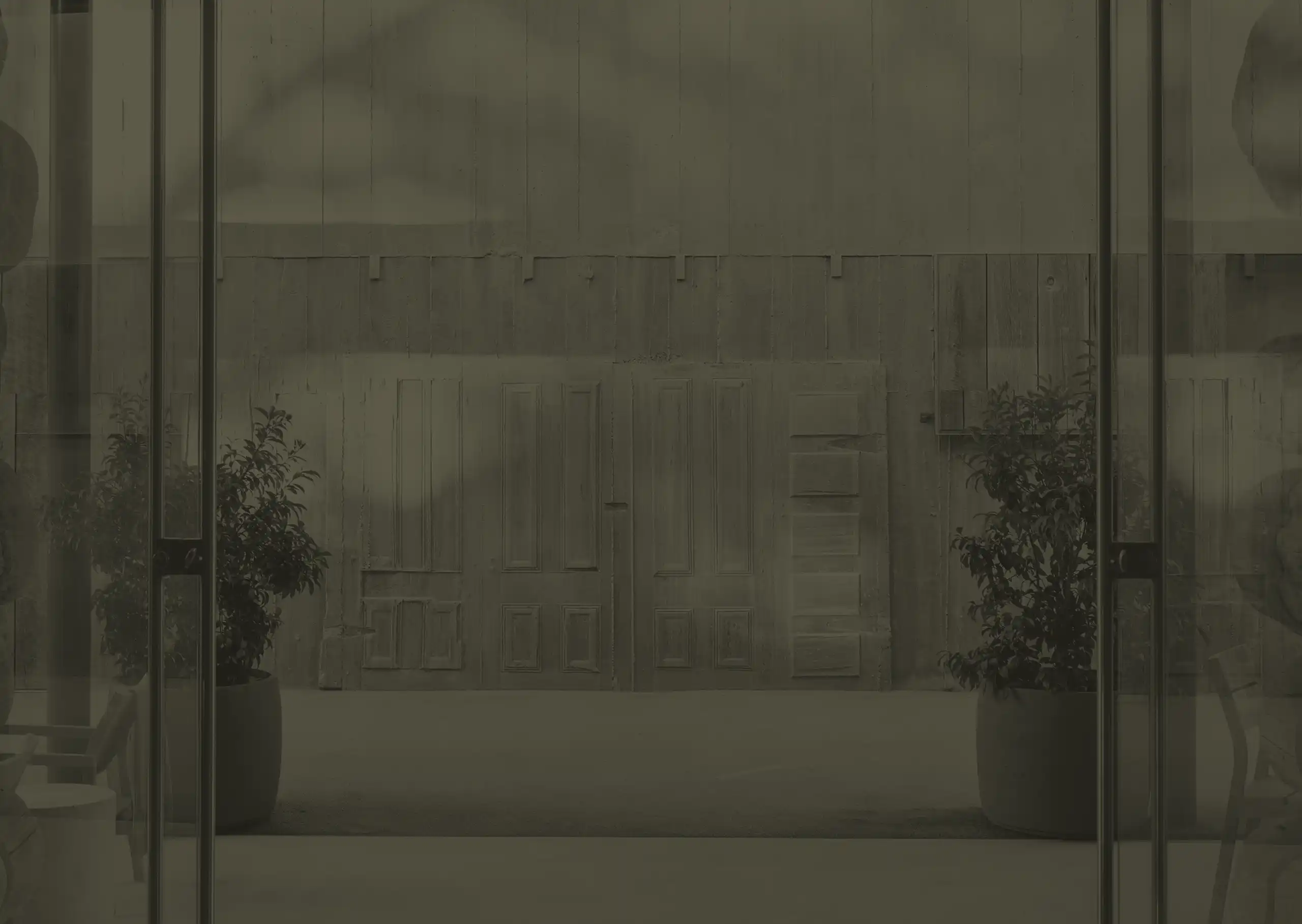 Two large potted plants flank a wall with multiple old wooden doors, viewed through glass doors, in a dimly lit, industrial-style space.