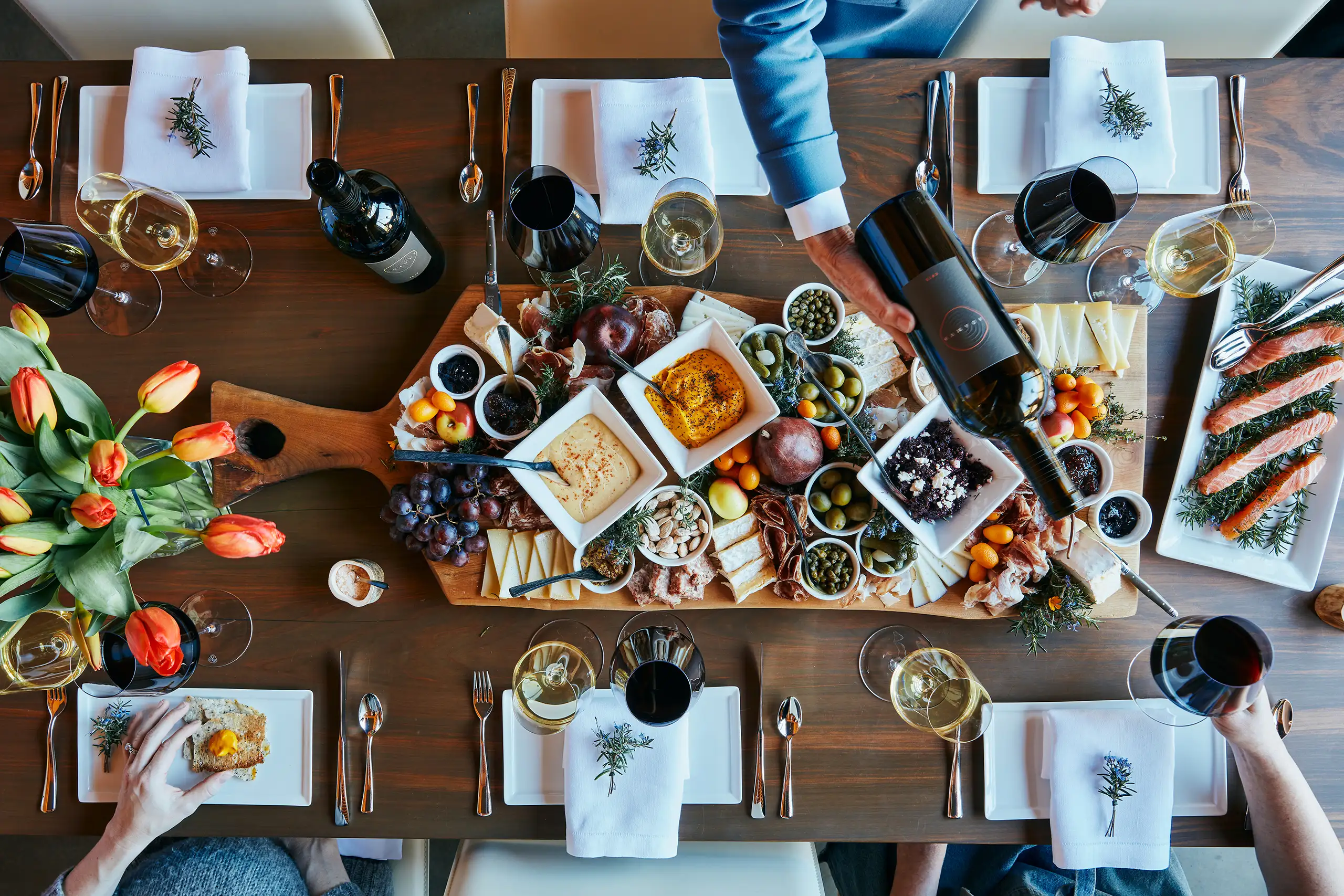 A wooden table set for a meal with wine, glasses, plates, flowers, and a large charcuterie board with various foods at the center. People serve themselves from the spread.