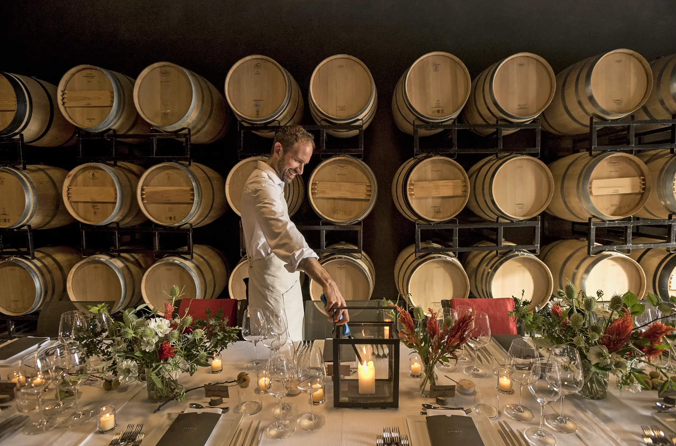 A man lights a candle on a set dining table in front of stacked wine barrels, with flowers, glassware, and menus arranged neatly.