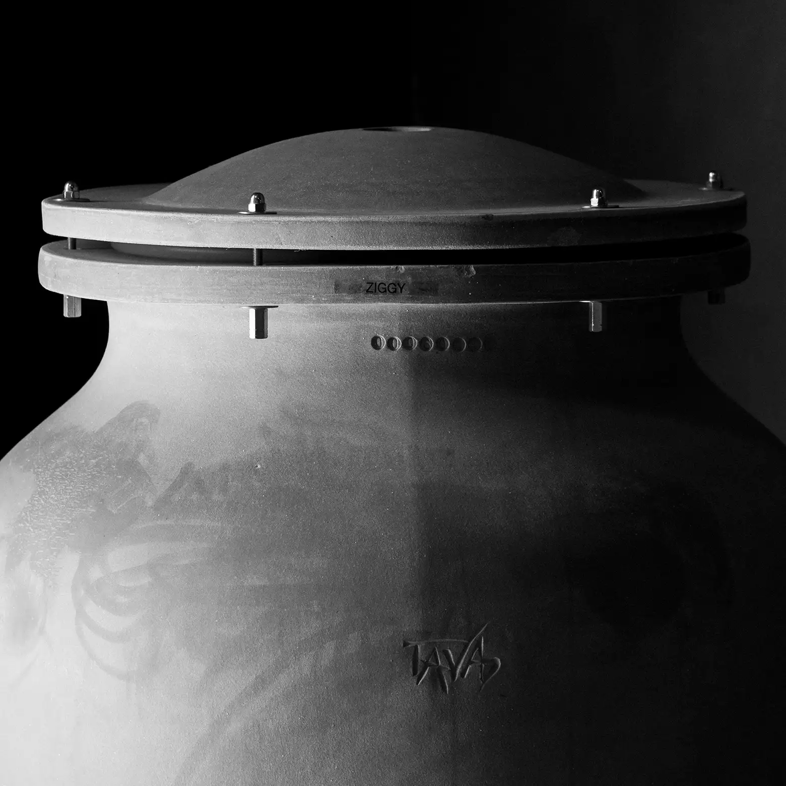 Black and white photo of a large, round ceramic grill with a domed lid labeled “ZIGGY” and “TAVA,” shown against a dark background.