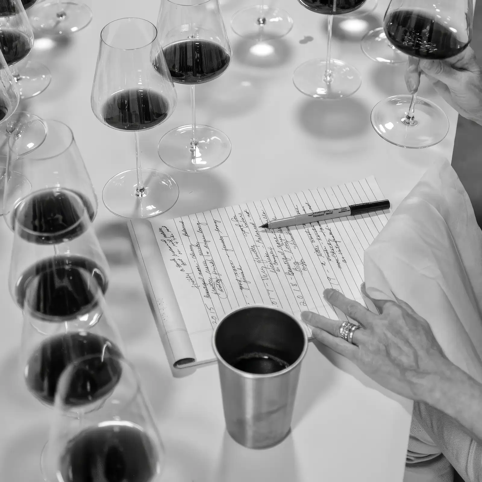 Several wine glasses with red wine are arranged on a table beside a notepad, pen, and a person’s hands taking notes.