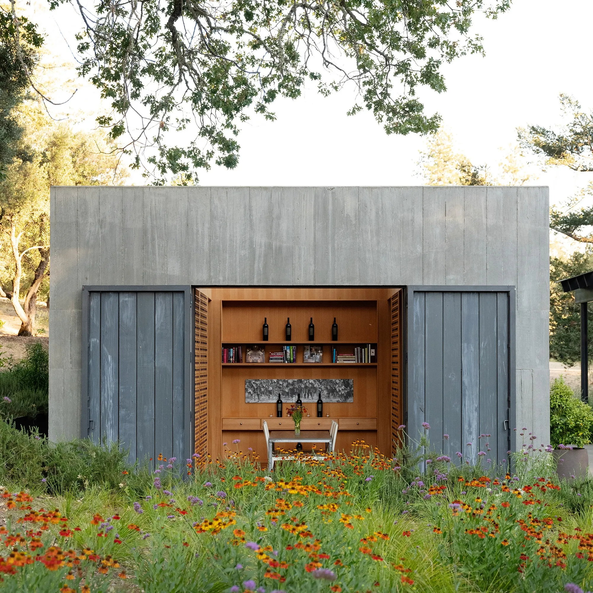 A modern concrete structure with open sliding doors reveals a wood-lined alcove featuring shelves of wine bottles and books, a small table, and a meadow of wildflowers in the foreground.